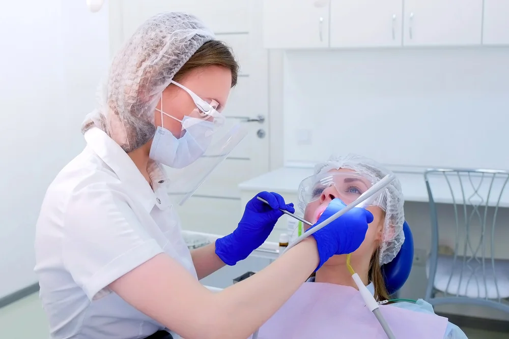 Dental professional wearing protective equipment uses dental instruments in a reclined patient’s mouth.
